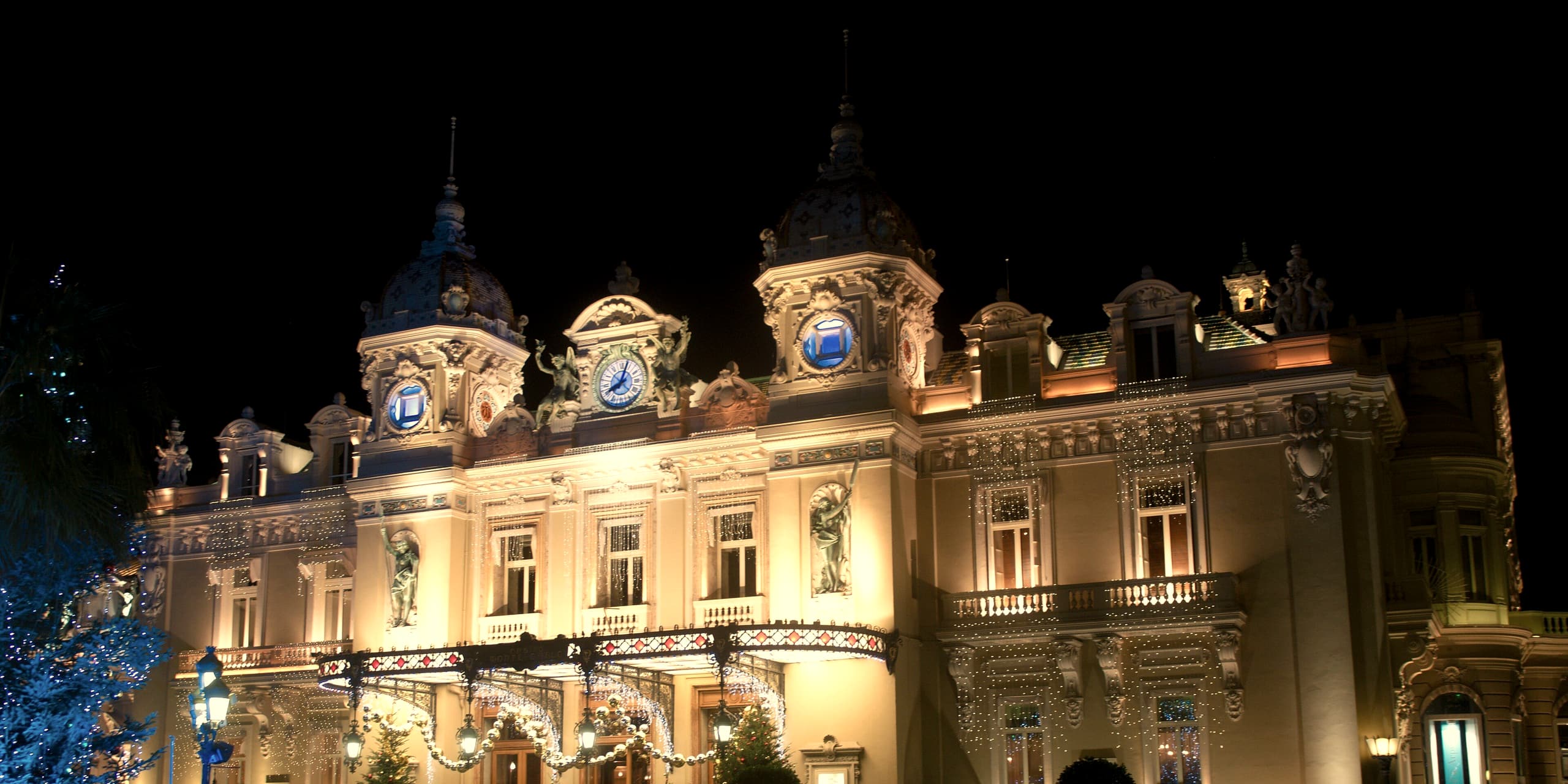 The Casino de Monte-Carlo at night, its domed towers, clocks and iron canopy lit warm against a black sky and strung with fine winter lights.