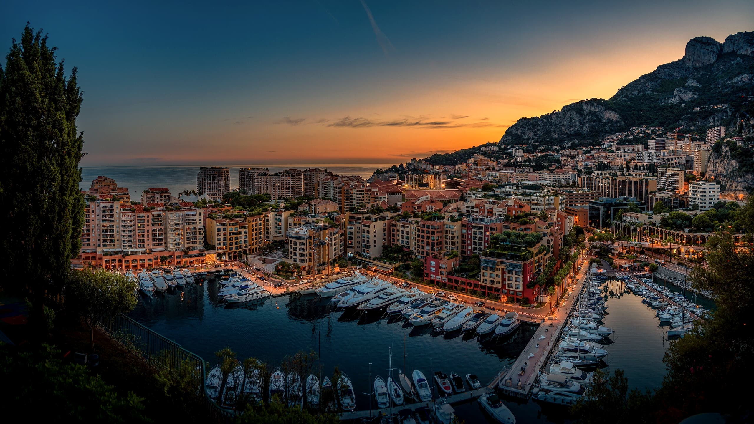 Fontvieille harbour at dusk, yachts moored below lit apartment buildings, the sea on the horizon and the mountain catching the last orange light.