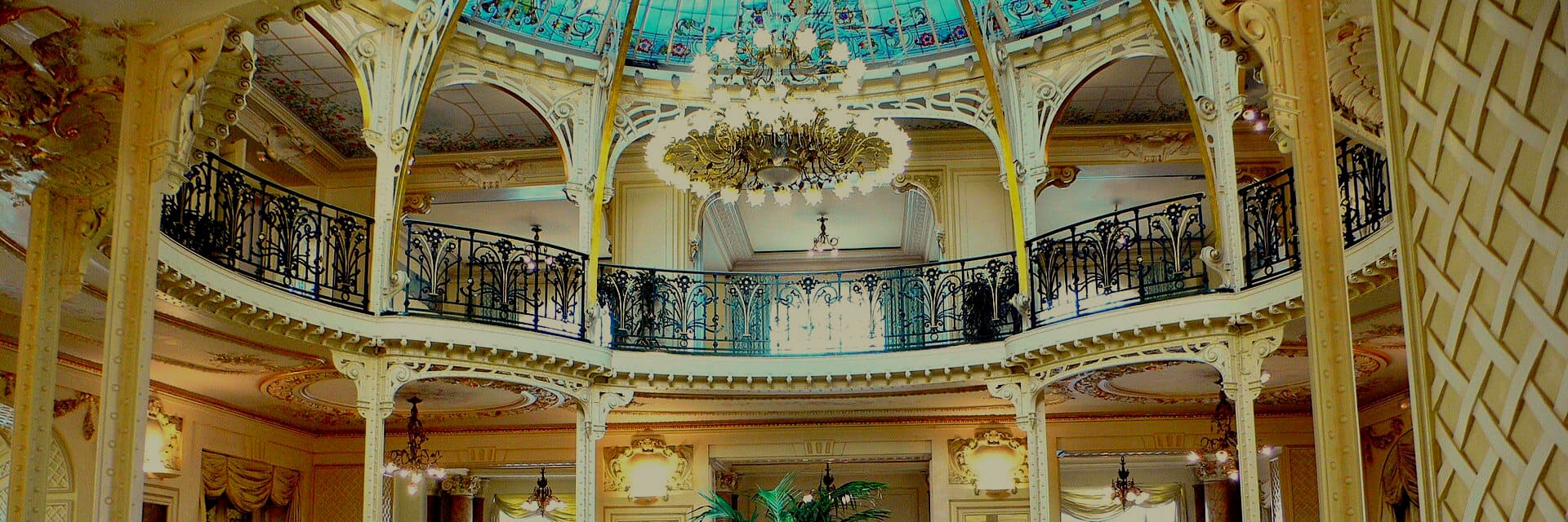A Belle Epoque winter garden beneath a stained glass dome, a chandelier hanging above a curved wrought iron balcony.