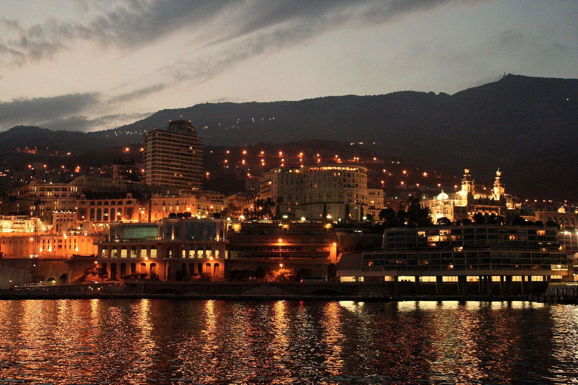 Monaco seen across the water at dusk, lit buildings along the shore reflected in the sea beneath dark mountains.