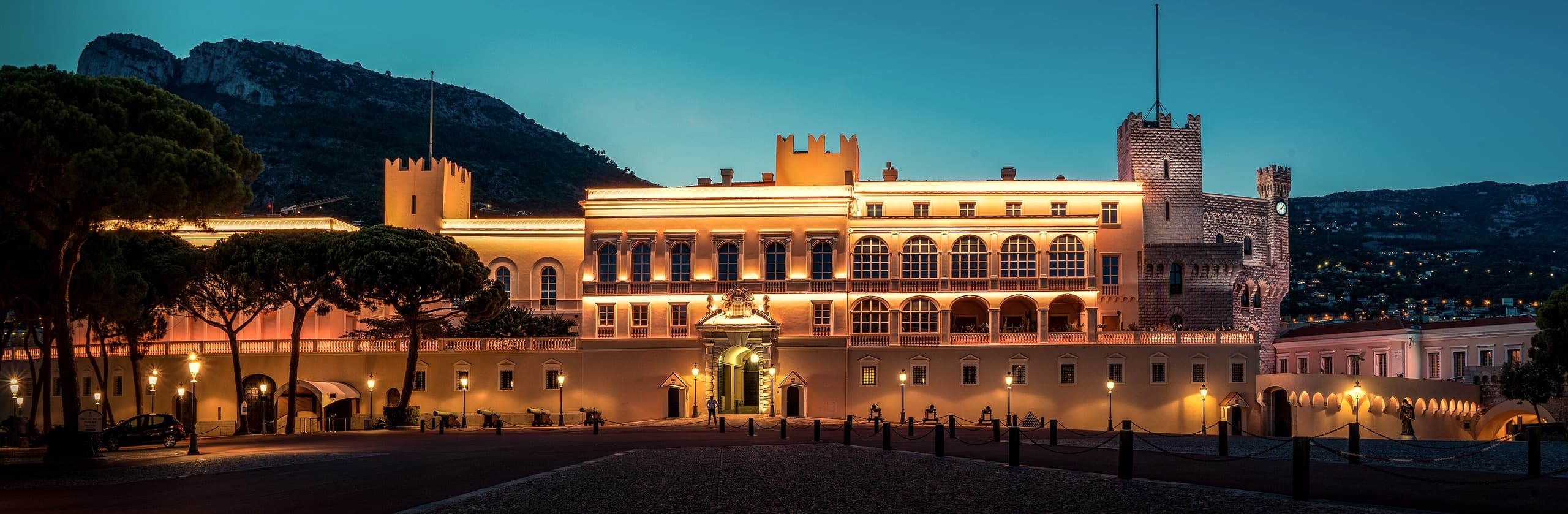 The square on the Rocher at blue hour, the long palace façade lit warm gold beneath umbrella pines and a deep blue sky.